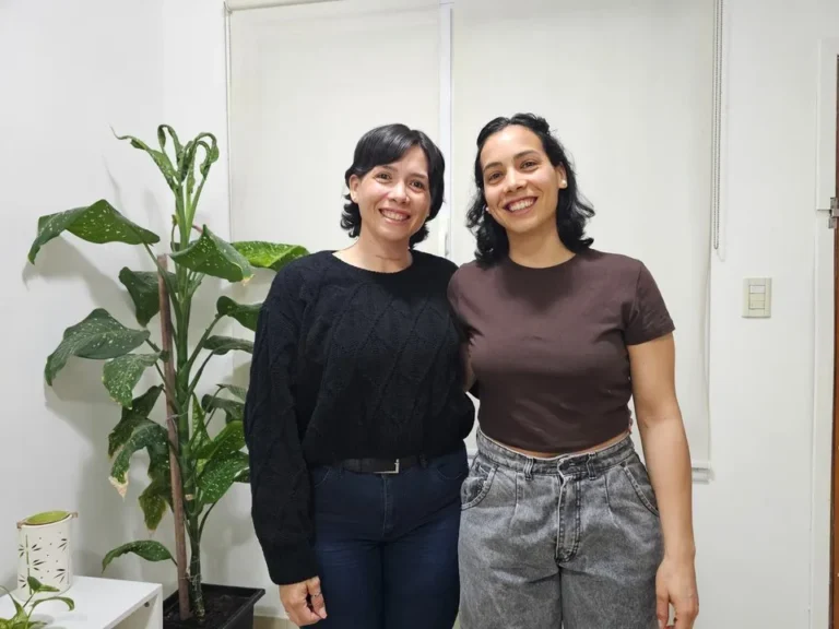 Dos hermanas sonriendo, una con cabello corto y otra con pañuelo en la cabeza, sentadas en un sillón.